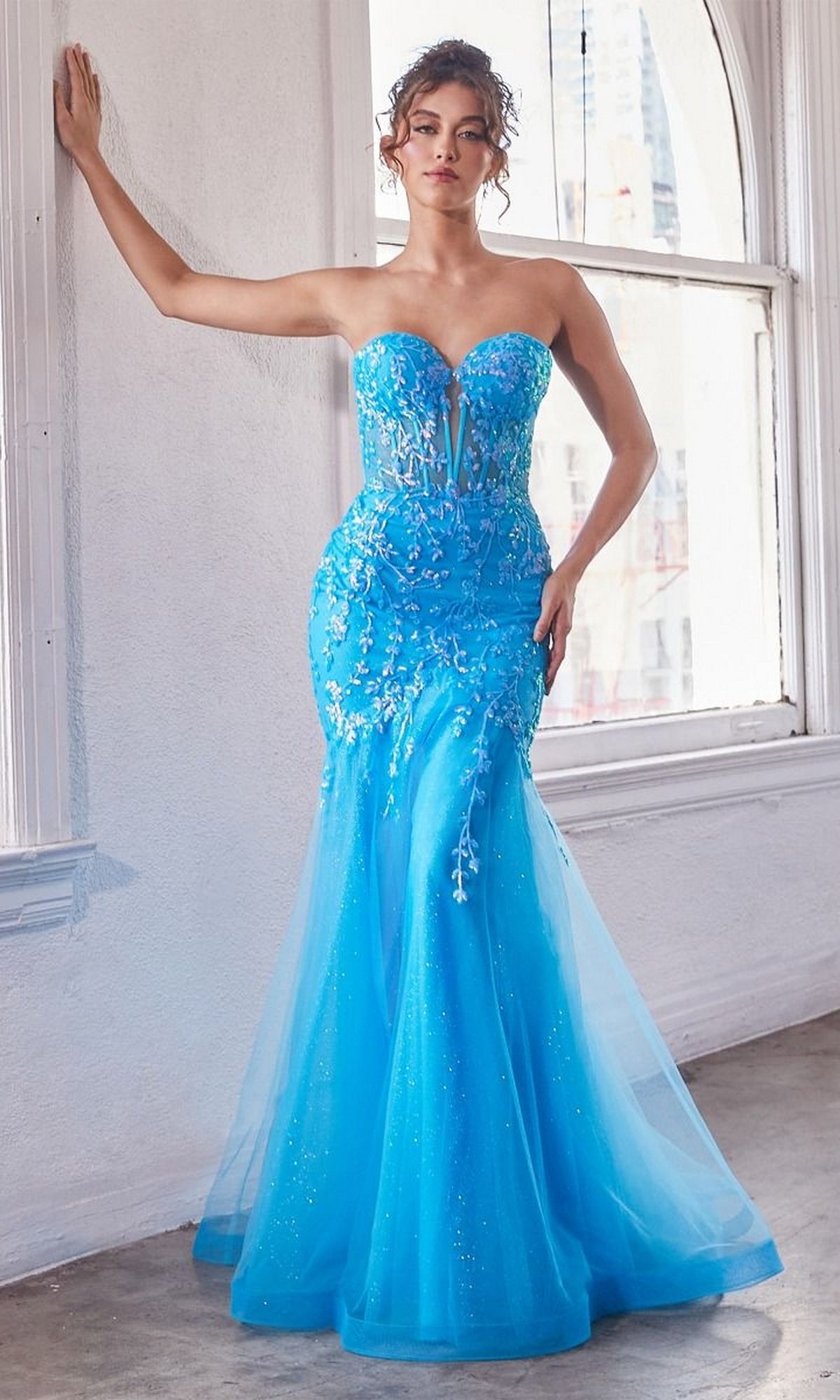 A woman stands by a window in the Strapless Sweetheart Long Mermaid Prom Dress CB139—a fitted, bright blue gown with floral appliqué and tulle skirt—ideal as an embroidered formal dress.